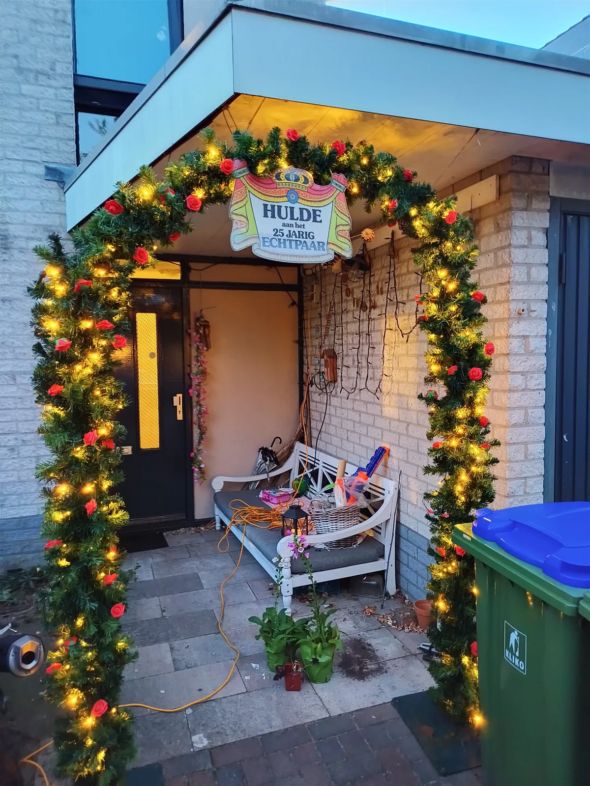 Groene trouwboog met rode kunstrozen, warme lampjes en een Hulde-bord bij een voordeur